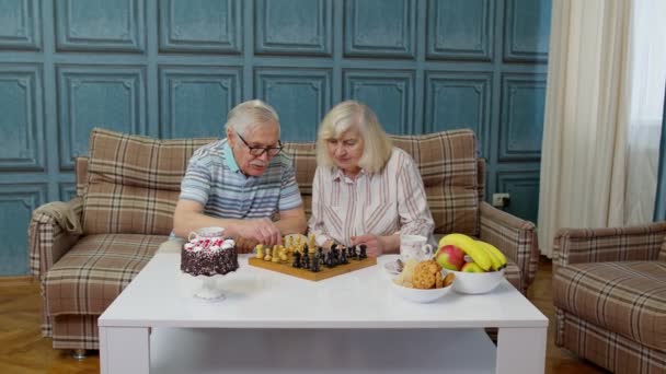 Пенсионерская пара разговаривает, пьет чай, играет в шахматы в современной гостиной вместе - Кадры, видео