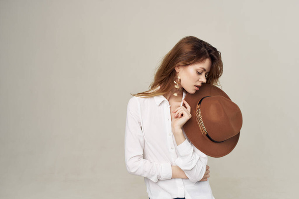 ładna kobieta trzyma kapelusz w jej ręce biały koszula moda styl życia beżowy tło - Zdjęcie, obraz