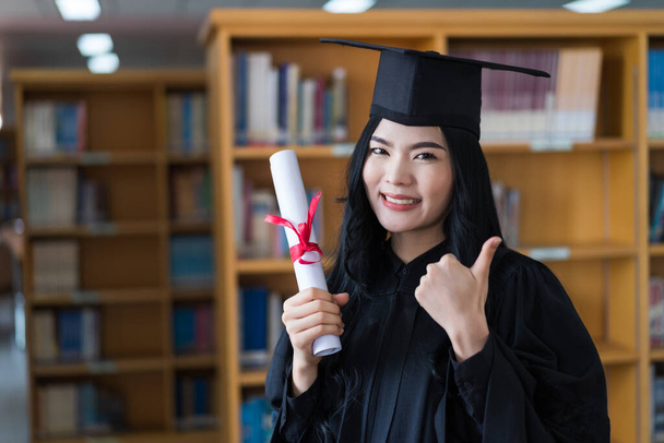 A young happy Asian woman university graduates in graduation gown and cap wears a face mask holds a degree certificate to celebrate her education achievement on the commencement day. Stock photo - Photo, Image