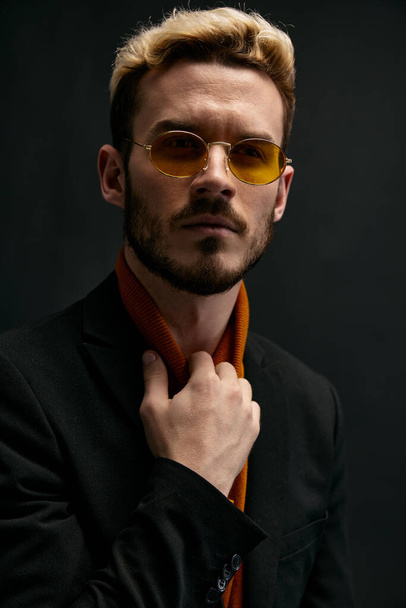 hombre con estilo con gafas chaqueta negra rubia suéter naranja fondo oscuro - Foto, Imagen