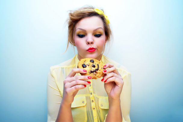 Jovem mulher atraente com cabelo vermelho e uma cabeça amarela arco vestindo uma blusa de chiffon amarelo está segurando um biscoito de chocolate sorridente chip e olhando para ele com um olhar indeciso
. - Foto, Imagem
