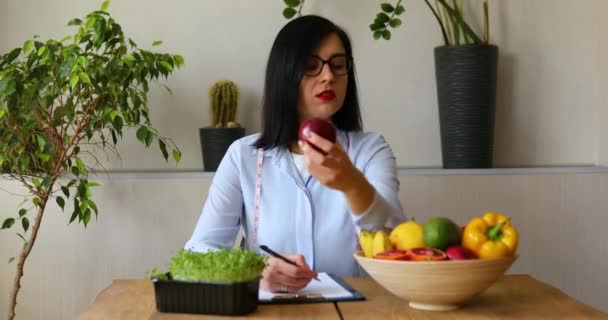 栄養士、事務所の栄養士の女性は、手にリンゴを保持し、健康的な野菜や果物、医療や食事の概念。彼女の机で働いている果物と女性栄養士. - 映像、動画