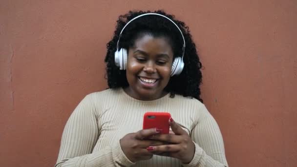 Glückliche Afroamerikanerin, die online mit ihrem Handy Nachrichten verschickt, während sie mit drahtlosen Kopfhörern Musik hört - Filmmaterial, Video