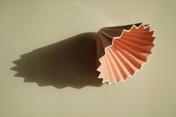 Růžové origami káva kapátko na bílém pozadí. Jasné sluneční světlo a výrazný geometrický stín - Fotografie, Obrázek