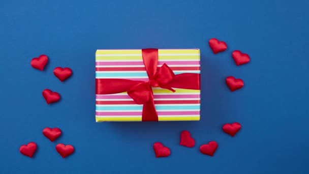 ingepakte kleurrijke geschenkdoos met rood lint en rode hartjes op blauwe achtergrond, bovenaanzicht. stop beweging animatie - Video