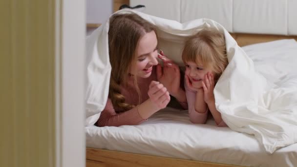 Happy family of mother with daughter lying on bed under duvet blanket, telling secrets to each other - Footage, Video
