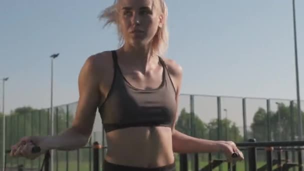 Low-angle medium shot of young attractive sportswoman in sport crop top jumping skipping rope outside during workout - Footage, Video