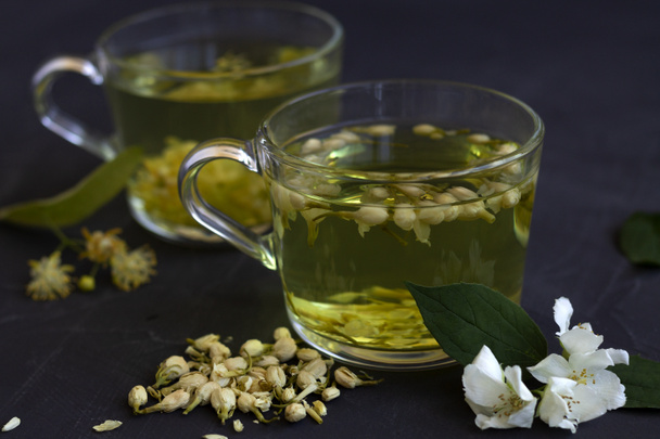 ジャスミン茶。黒テーブルの上にリンデンの花の熱いハーブティー2杯。健康的なライフスタイル。黒を基調とした緑茶のグラス。ホットドリンク. - 写真・画像