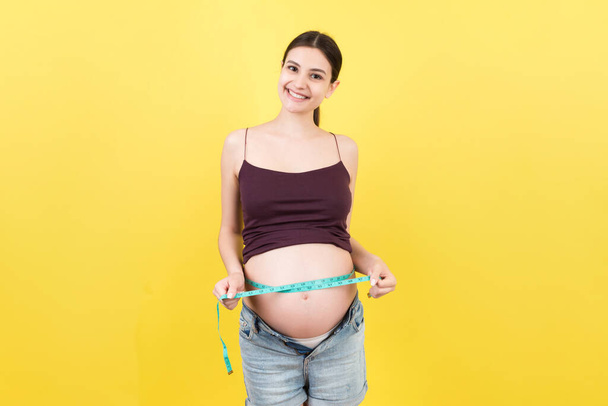 Imagen recortada de la madre embarazada en jeans desabrochados midiendo su vientre embarazada con una cinta métrica en un fondo colorido. Medición del centímetro. - Foto, imagen