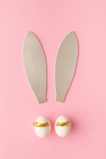 Rabbit ears made of silver foam. Two white eggs with golden lashes. Bunny face concept. Easter pink background. - Foto, Imagen