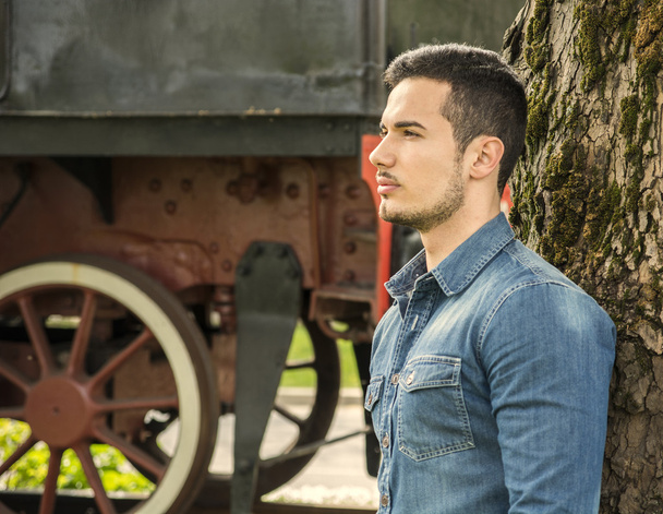 Profile of young man in denim shirt near old train, against tree - Foto, immagini