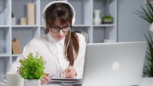 Młoda kobieta w słuchawkach pisze w notesie patrząc na laptopa. Uczennica jest szkolona zdalnie za pomocą komputera. Webinarium online rozmowy wideo. - Materiał filmowy, wideo