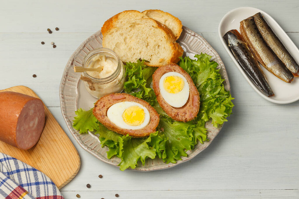 Σκωτσέζικα αυγά σε ένα πιάτο με μαρούλι, φρυγανισμένο ψωμί και σάλτσα. Σε ανοιχτό γκρι ξύλινο φόντο με ζαμπόν και σαρδέλες. Κοντινό πλάνο, αντίγραφο χώρου. Βρετανικό πρωινό, μεσημεριανό - Φωτογραφία, εικόνα