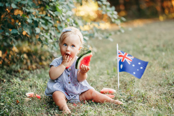 Roztomilý rozkošný kavkazský dítě dívka jíst zralé meloun s australskou vlajkou na zemi. Vtipné dítě sedí v parku s čerstvým ovocem slaví Austrálie Den dovolené venku. - Fotografie, Obrázek