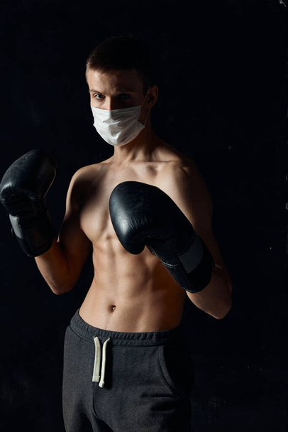 γάντια πυγμαχίας αθλητή σε μαύρο φόντο ιατρική μάσκα στο πρόσωπο γυμνό κορμό - Φωτογραφία, εικόνα