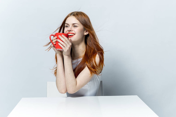mujer pelirroja sentada a la mesa sosteniendo una taza con un fondo claro de bebida - Foto, imagen