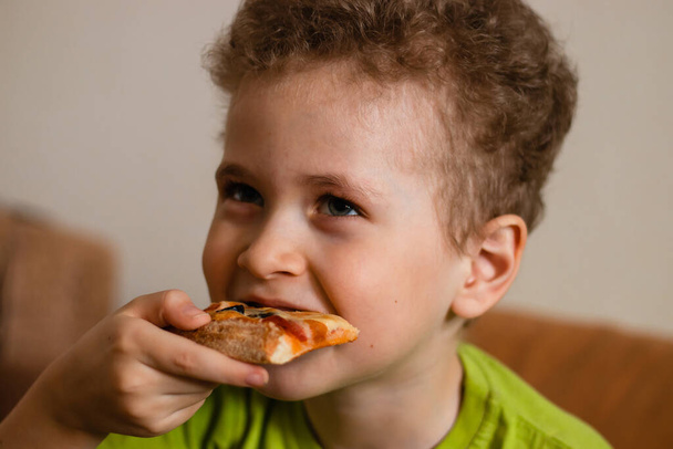 Un chico con el pelo rizado y una camiseta verde toma un bocado de pizza. El niño come en casa - Foto, imagen