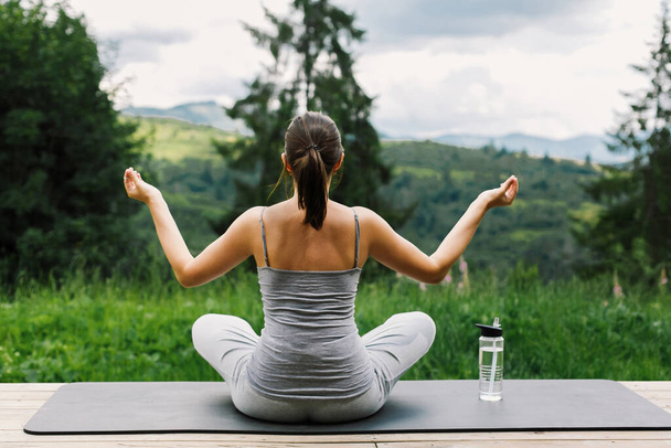 Młoda, sprawna kobieta praktykująca medytację na macie do jogi na tle słonecznych gór. Trening na świeżym powietrzu i trening. Zdrowy styl życia. Sportowy Kobieta robi jogę na drewnianym tarasie wśród drzew - Zdjęcie, obraz
