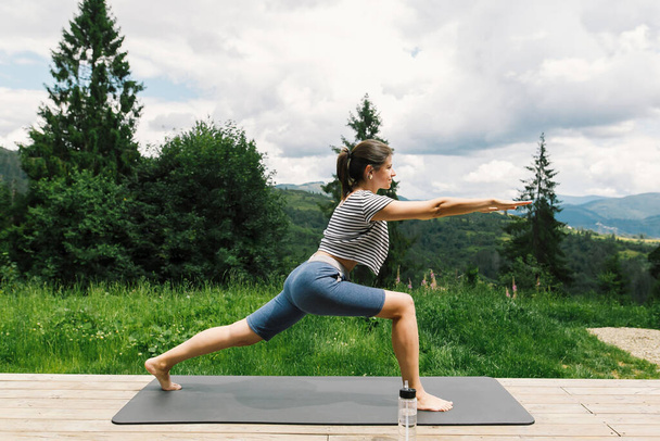Giovane donna in forma stretching o facendo squat su tappetino yoga sullo sfondo di colline di montagna soleggiate. Allenamento e allenamento all'aperto. Donna casual sulla terrazza in legno tra gli alberi, stile di vita sano - Foto, immagini
