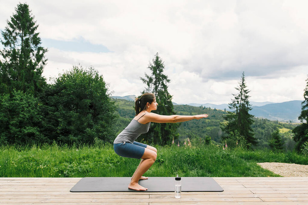 Giovane donna in forma stretching o facendo squat su tappetino yoga sullo sfondo di colline di montagna soleggiate. Allenamento e allenamento all'aperto. Donna casual sulla terrazza in legno tra gli alberi, stile di vita sano - Foto, immagini