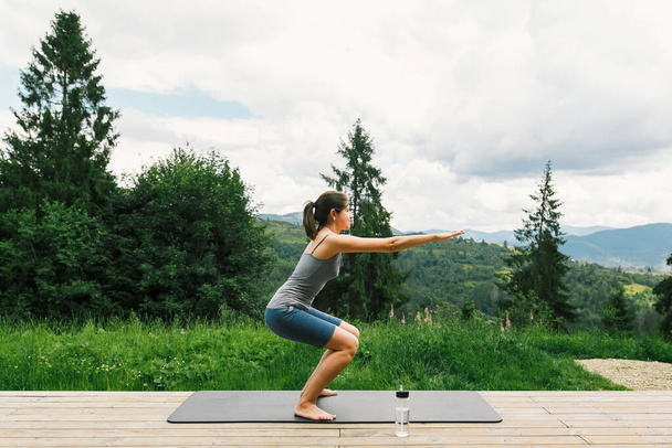 Junge, fitte Frau dehnt sich oder macht Kniebeugen auf einer Yogamatte vor dem Hintergrund sonniger Berghügel. Outdoor-Training und -Training. Lässiges Weibchen auf Holzterrasse zwischen Bäumen, gesunder Lebensstil - Foto, Bild