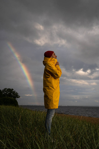 Man in gele regenjas draagt rode hoed op het strand bij regenachtig weer, kijkt naar dramatische bewolkte lucht en zee, regenboog op de achtergrond. Herfstseizoen. - Foto, afbeelding