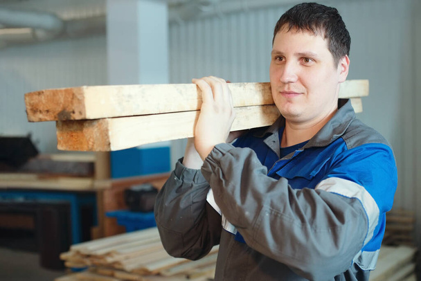 Pracownik tartaku w kombinezonie nosi drewniane pręty na ramieniu. Produkcja i dostawa materiałów budowlanych. - Zdjęcie, obraz