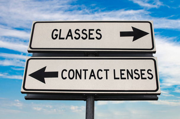 Szemüveg kontra kontaktlencse útjelzés két nyíllal a kék ég hátterében. Fehér, két utcás tábla, nyilakkal a fémrúdon. Kétirányú jelzőtábla szöveggel. - Fotó, kép
