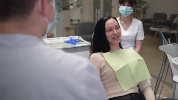 Uma jovem caucasiana na consulta médica senta-se em uma cadeira dentária e se diverte conversando com o médico. Saúde dentária. - Filmagem, Vídeo