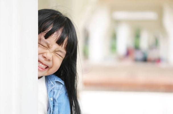 Asian children cute or kid girl wear jeans for fashion and fun playing hide and seek with sneaked or dodge behind post and close eye with smile white teeth in pavilion at park garden on relax holiday - Photo, Image