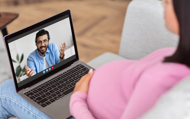 Communicatieconcept. Achteraanzicht van zwanger meisje het maken van video bellen op laptop thuis, chatten met opgewonden echtgenoot - Foto, afbeelding