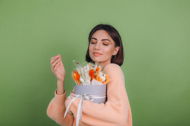 Молода жінка в повсякденному персиковому светрі ізольована на зеленому оливковому фоні тримає помаранчеву білу квіткову коробку композицію з бавовняних квітів, гіпсофіли, пшениці та лагуни для подарунка щасливе здивоване обличчя
 - Фото, зображення