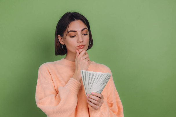 Mladá žena v ležérní broskvový svetr izolované na zeleném olivovém pozadí štěstí drží fanoušek 100 dolarů bankovky přemýšlivý počítání hotovosti - Fotografie, Obrázek