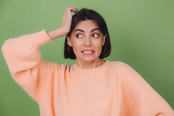 Молодая стильная женщина в обычном персиковом свитере и оранжевых очках изолированы на зеленом оливковом фоне озабочены положить руку на голову смотреть в сторону копировать пространство - Фото, изображение