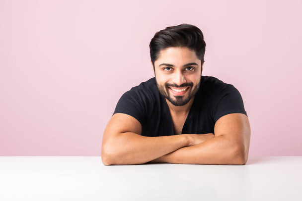 Portrét šťastné indické asijské vousatý muž sedí u stolu nebo stolu nebo platformy proti růžové pozadí při pohledu do kamery - Fotografie, Obrázek