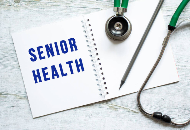 SENIOR HEALTH is written in a notebook on a light wooden table next to pencil and a stethoscope. Medical concept - Photo, Image