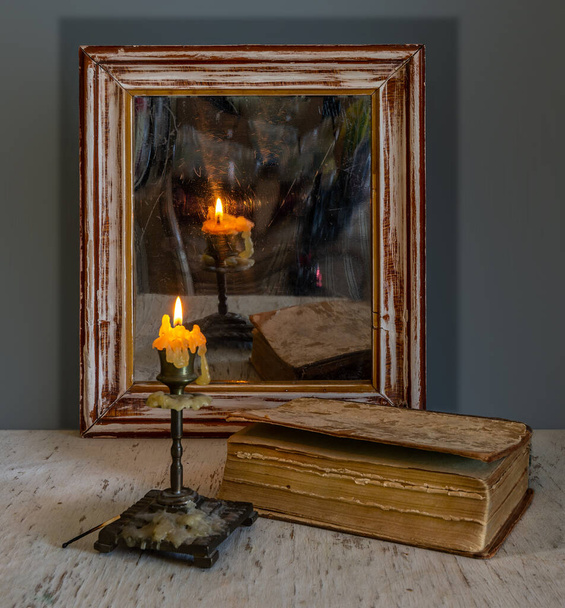 Ένα αναμμένο κερί αντανακλάται σε έναν παλιό καθρέφτη σε ένα ξύλινο πλαίσιο και φωτίζει τα βιβλία στο τραπέζι. Εσοδεία. - Φωτογραφία, εικόνα