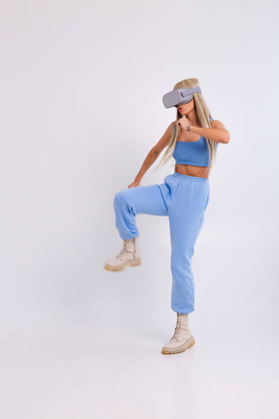 Studio photo of a young attractive woman in a warm blue fashionable suit wearing virtual reality glasses on a white background plays a boxing fight - Photo, Image