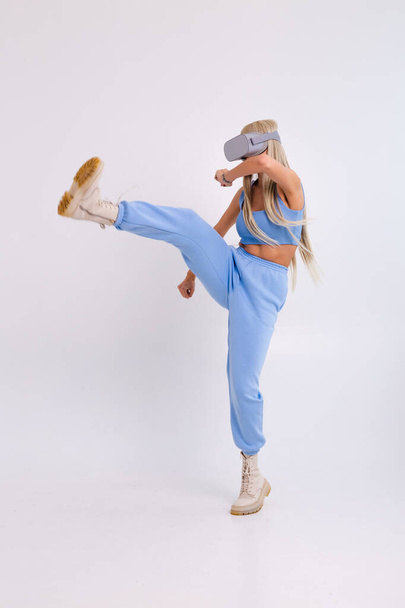 Studio photo of a young attractive woman in a warm blue fashionable suit wearing virtual reality glasses on a white background plays a boxing fight - Photo, Image
