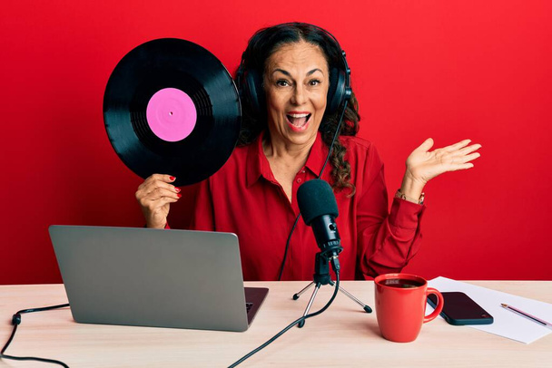 Mooie middelbare leeftijd vrouw werken op radio studio holding vinyl schijf vieren prestatie met een vrolijke glimlach en winnaar uitdrukking met opgeheven hand  - Foto, afbeelding