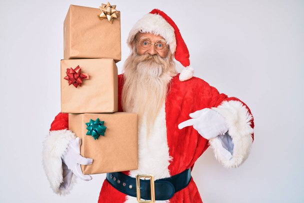 Γέρος ηλικιωμένος άνδρας με γκρίζα μαλλιά και μακριά γενειάδα φορώντας κοστούμι Santa Claus κρατώντας δώρα δείχνοντας το δάχτυλο σε έναν εαυτό χαμογελαστός ευτυχισμένος και περήφανος  - Φωτογραφία, εικόνα