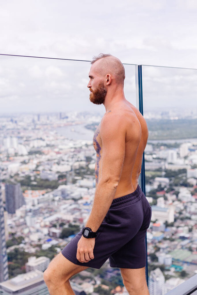 tattooed strong man topless, with watch, sits on chair on high floor - 写真・画像