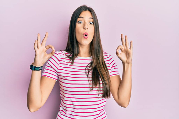 Young brunette woman wearing casual clothes over pink background looking surprised and shocked doing ok approval symbol with fingers. crazy expression  - Zdjęcie, obraz