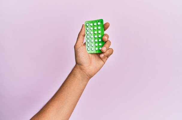 Joven mano hispana sosteniendo píldoras anticonceptivas sobre fondo rosa aislado. - Foto, Imagen