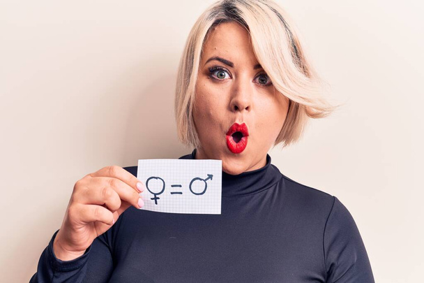 Blonde plus size woman asking for sex equality holding paper with gender equal message scared and amazed with open mouth for surprise, disbelief face - Photo, Image