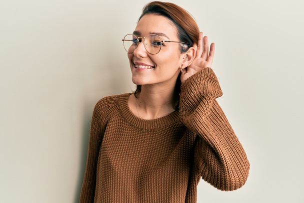 Junge kaukasische Frau in lässiger Kleidung und Brille, die lächelnd mit der Hand über dem Ohr Gerüchten oder Klatsch lauscht. Konzept für Gehörlosigkeit.  - Foto, Bild
