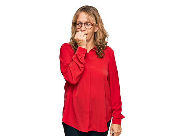 中世のブロンドの女性は、赤い背景にカジュアルなシャツを着て、口の中で爪をかむ上で手で強調し、緊張している。不安の問題.  - 写真・画像