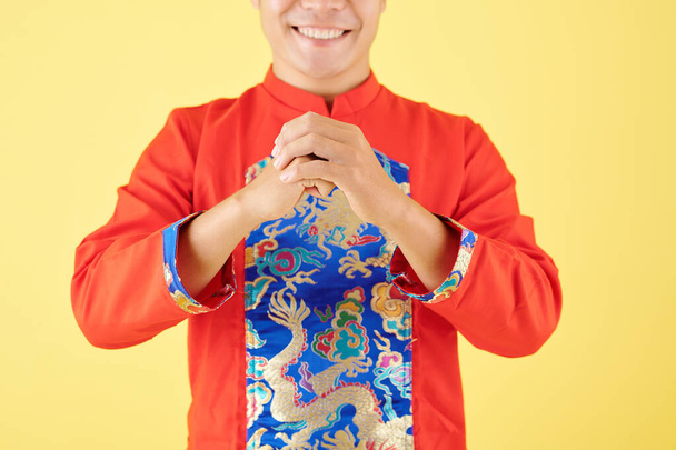 Oříznutý obraz veselého mladého Vietnamce v tradičním kostýmu, jak dělá gesto na pozdrav, izolovaný na žluté - Fotografie, Obrázek