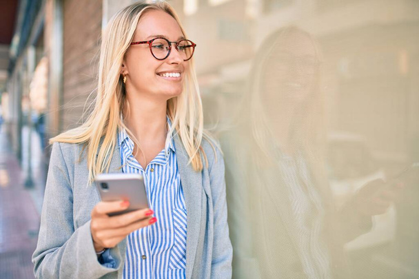 Νεαρή ξανθιά επιχειρηματίας χαμογελώντας ευτυχισμένη χρησιμοποιώντας smartphone στην πόλη. - Φωτογραφία, εικόνα