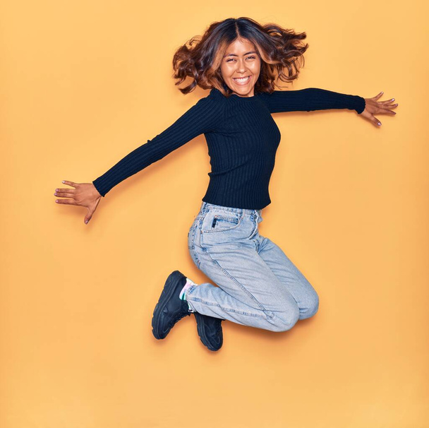 Young beautiful latin woman smiling happy. Jumping with smile on face and arms opened over isolated yellow background - Fotografie, Obrázek